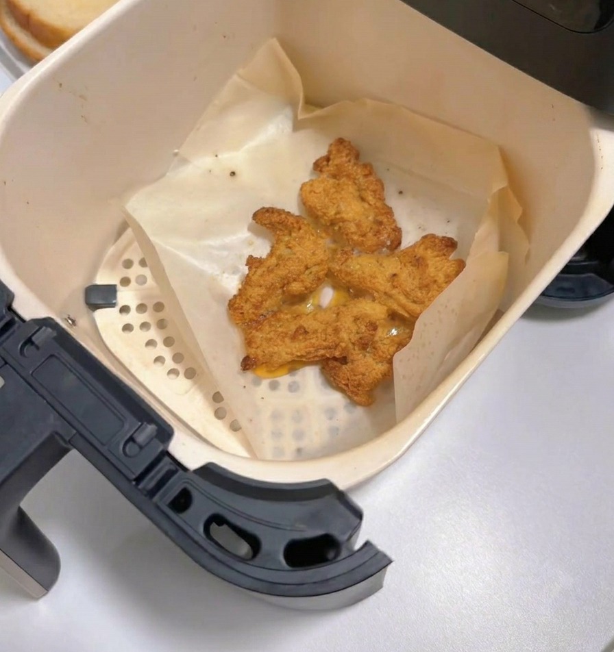 Cestello della friggitrice ad aria estratto che mostra pezzi di pollo fritto dorati e croccanti sulla carta forno.