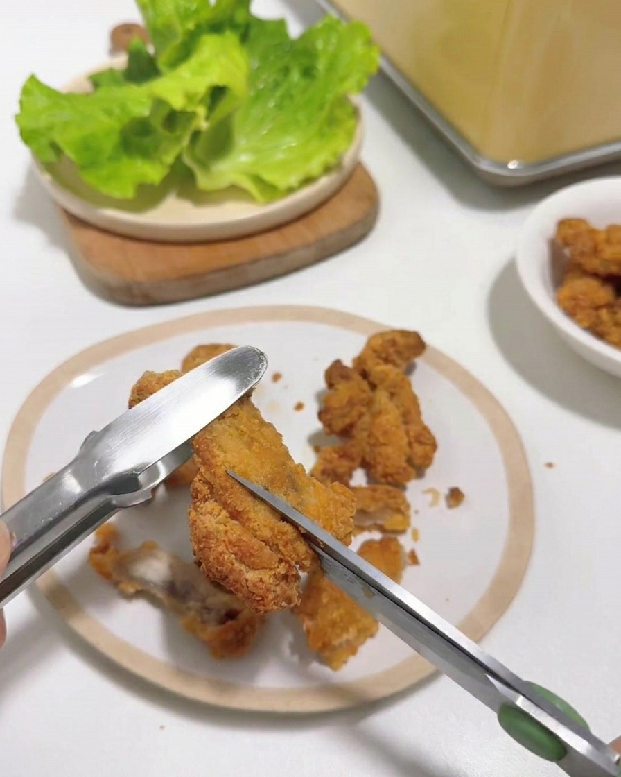 Primo piano di forbici da cucina che tagliano un pezzo di pollo fritto caldo tenuto con delle pinze su un piatto bianco.