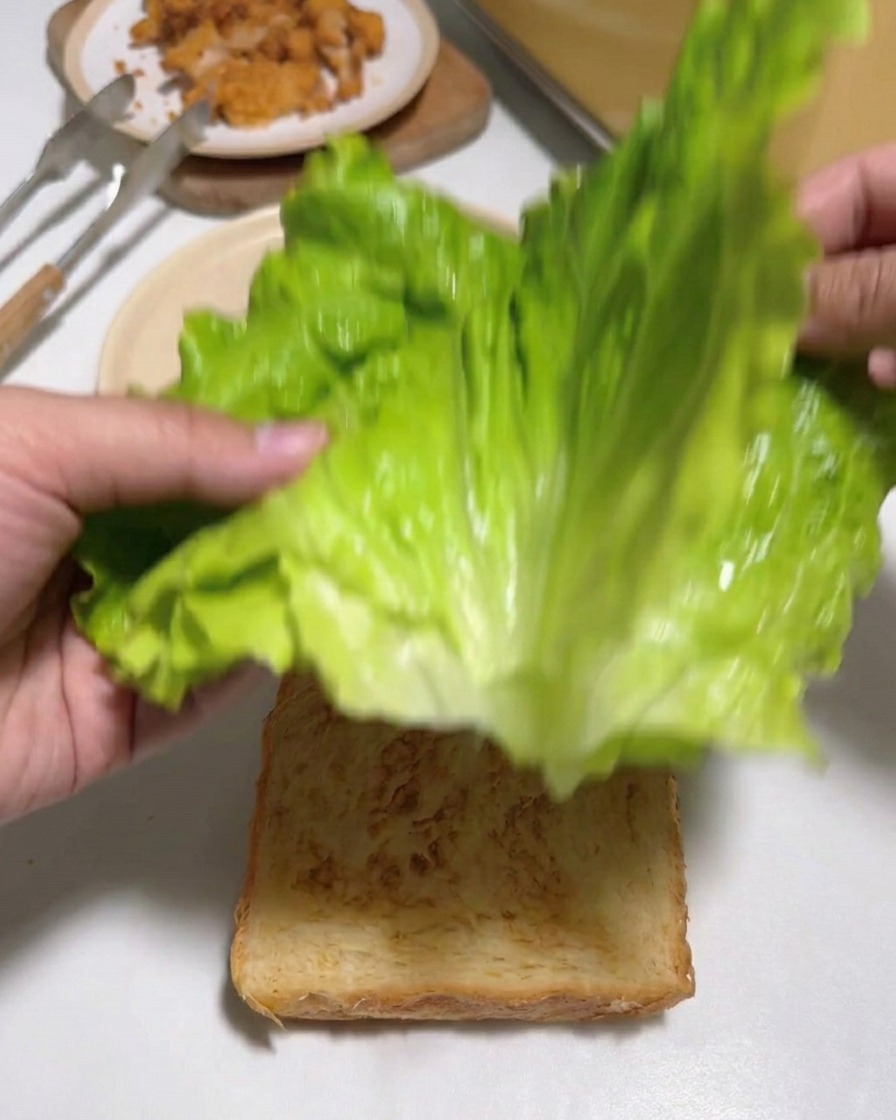 Mani che posizionano grandi foglie di lattuga verde fresca su una fetta di pane bianco tostato.
