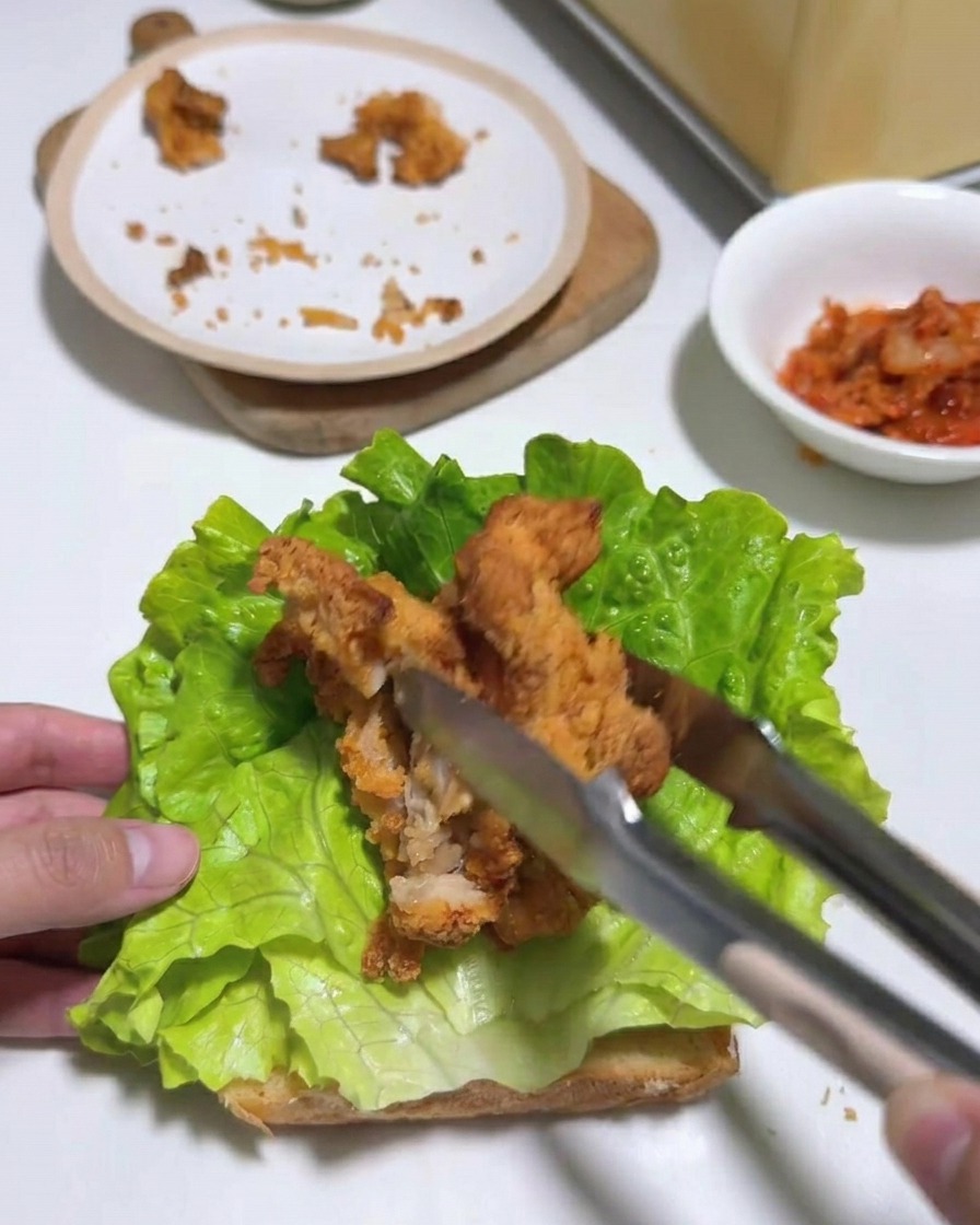 Pinze che posizionano diverse strisce di pollo fritto su uno strato di lattuga su pane tostato.