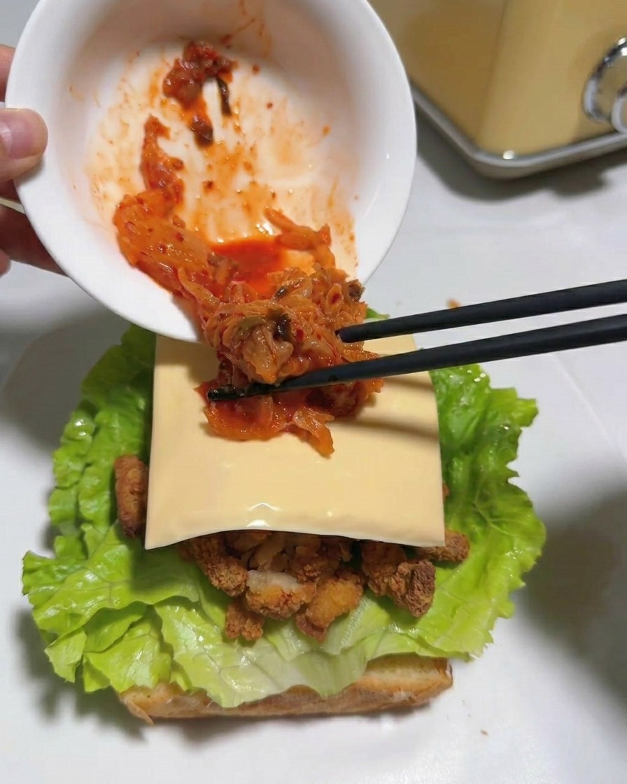 Bacchette che trasferiscono del kimchi tritato rosso acceso da una ciotola bianca su una fetta di formaggio in un sandwich.