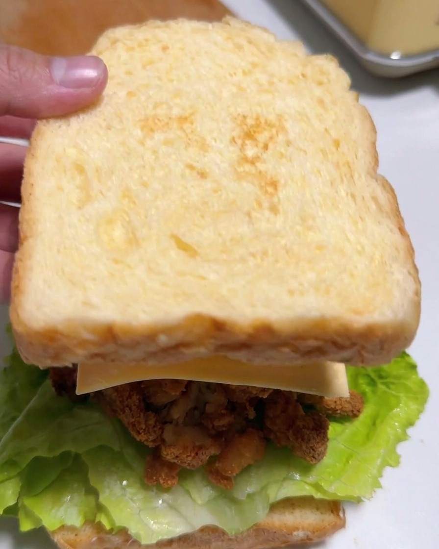 Una mano che posiziona una fetta di pane leggermente tostata sopra un sandwich farcito con lattuga, pollo, formaggio e kimchi.