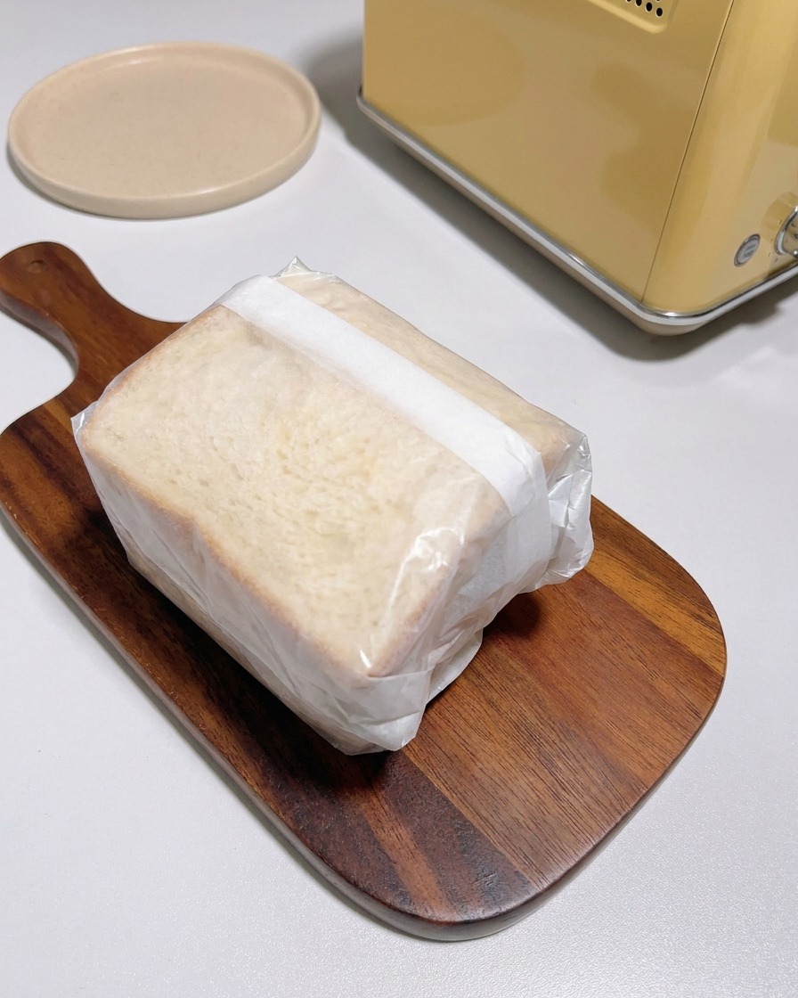 Un sandwich spesso avvolto strettamente in carta forno bianca traslucida su un tagliere di legno accanto a un tostapane.