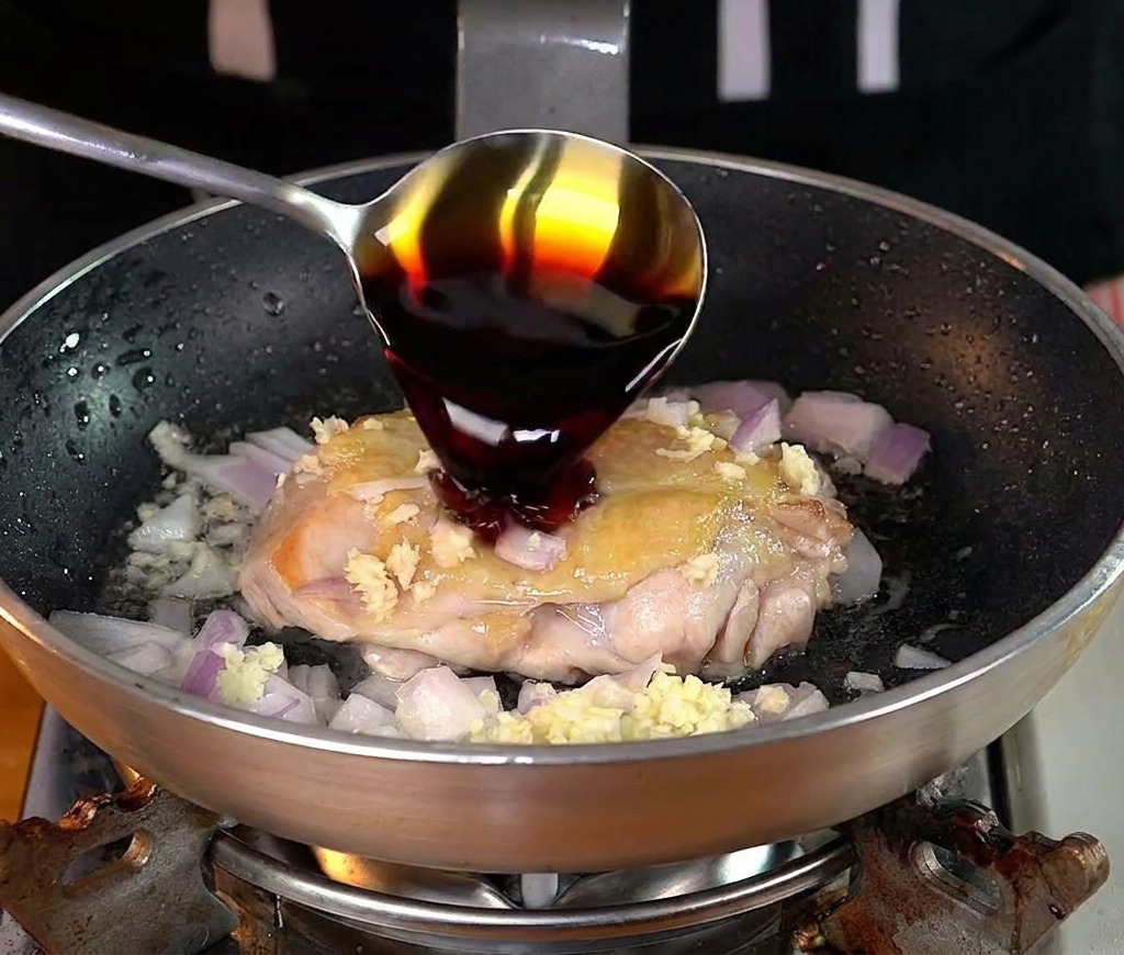 Un cucchiaio versa salsa di soia scura su una sovracoscia di pollo in padella.