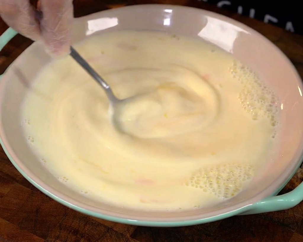 Un cucchiaio mescola uova e latte in una ciotola creando un vortice giallo chiaro.