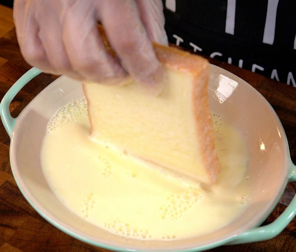 Una mano immerge una fetta spessa di pane bianco nel composto di uova e latte.