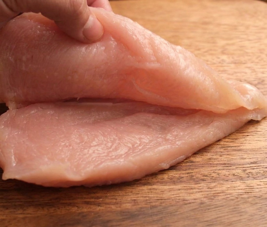 Una mano che taglia orizzontalmente un petto di pollo crudo in due pezzi sottili su un tagliere di legno.