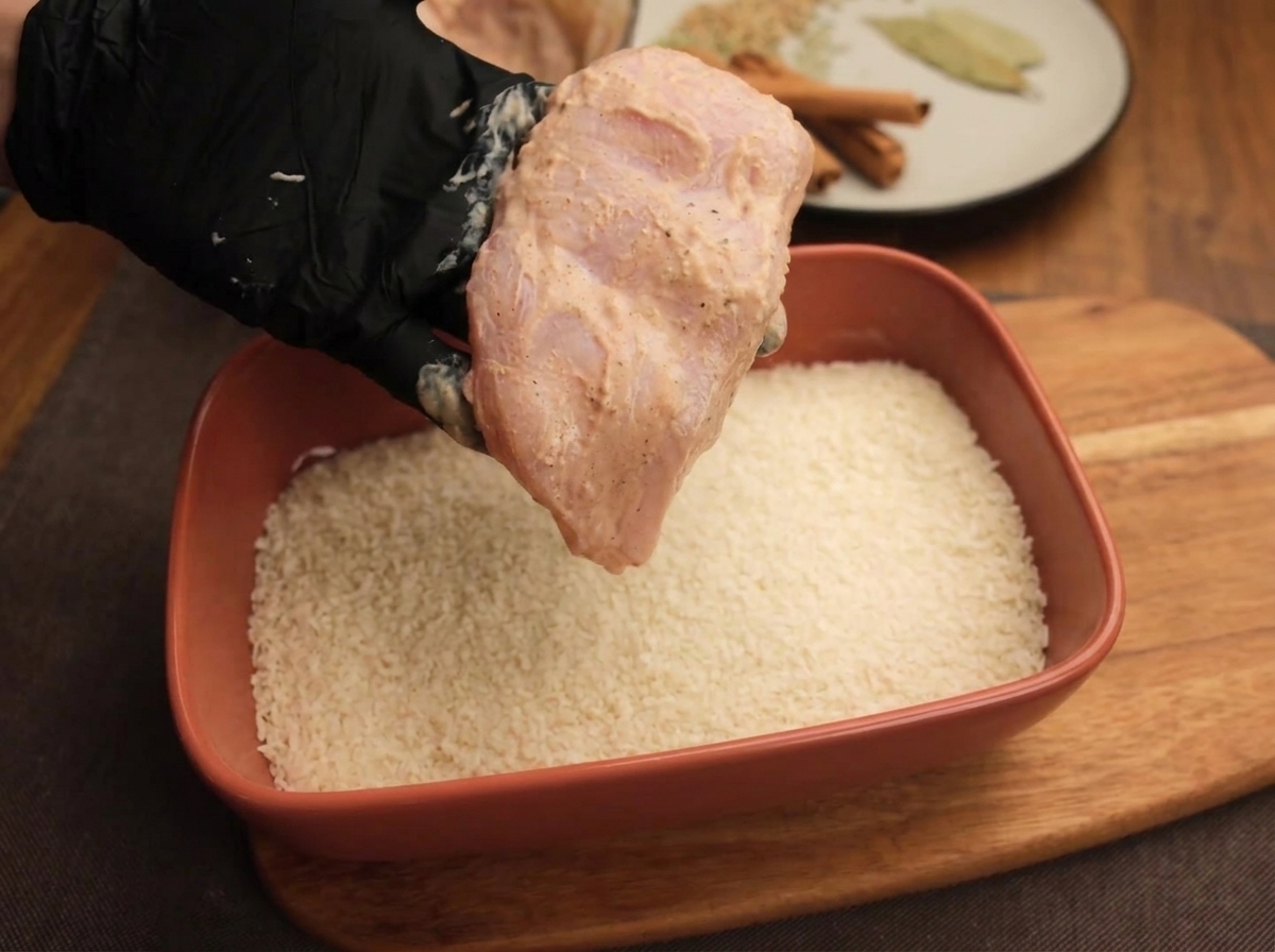 Mano guantata che tiene una fettina di pollo marinata sopra un vassoio di pangrattato bianco.