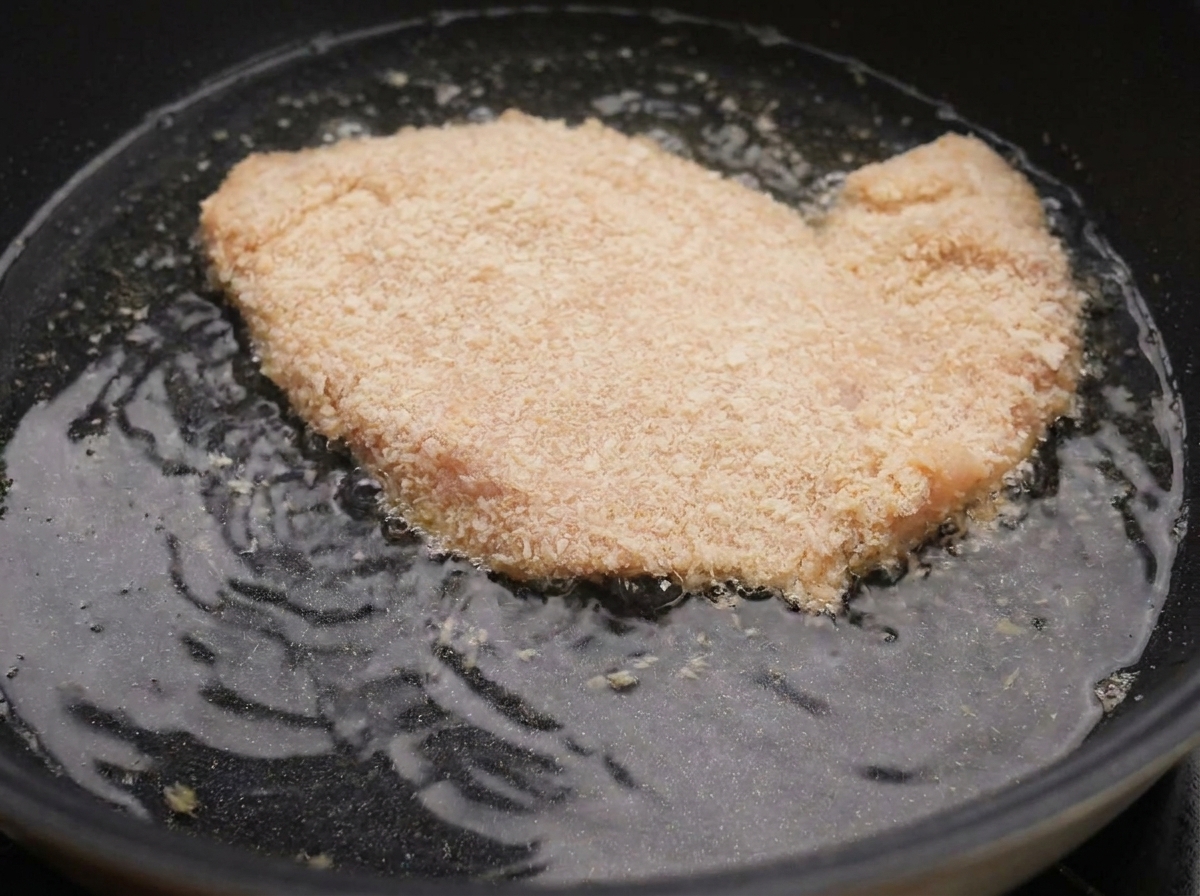 Petto di pollo impanato che sfrigola nellolio bollente allinterno di una padella nera.