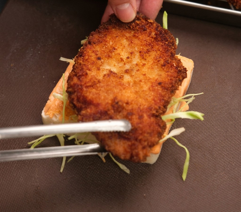 Mano e pinze che posizionano il pollo fritto su una fetta di pane con salsa e cavolo.