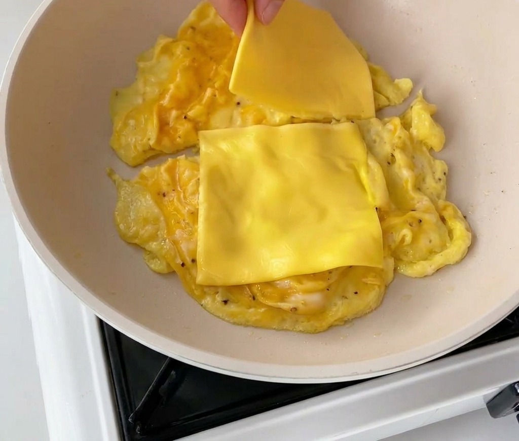 Una mano che adagia due fette di formaggio cheddar sulle uova strapazzate in padella.
