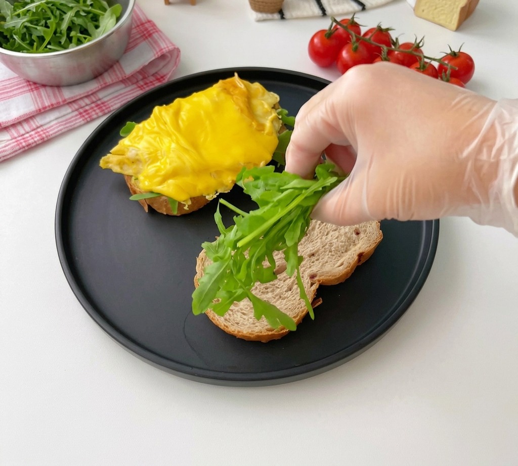 Una mano che mette foglie di rucola su una fetta di pane tostato su un piatto nero.