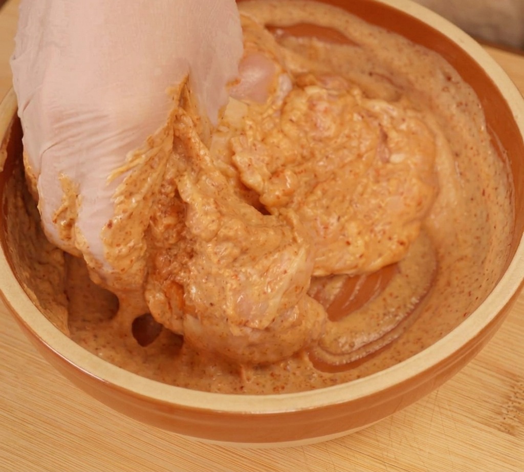 Una mano con guanto che massaggia il pollo nella marinata di yogurt di colore arancione.