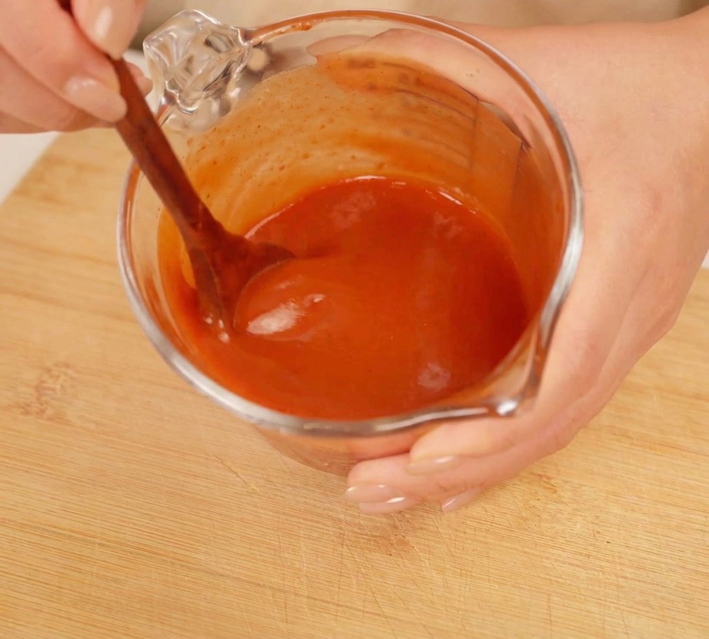 Mani che mescolano una salsa rossa densa in un misurino di vetro con un cucchiaio di legno.