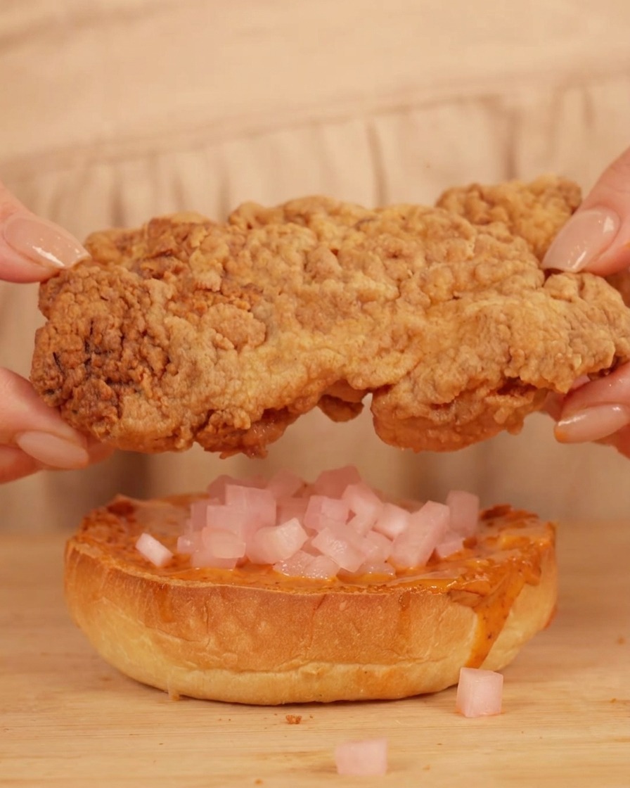 Mani che posizionano il pollo sopra il panino con salsa e cubetti di ravanello rosa.