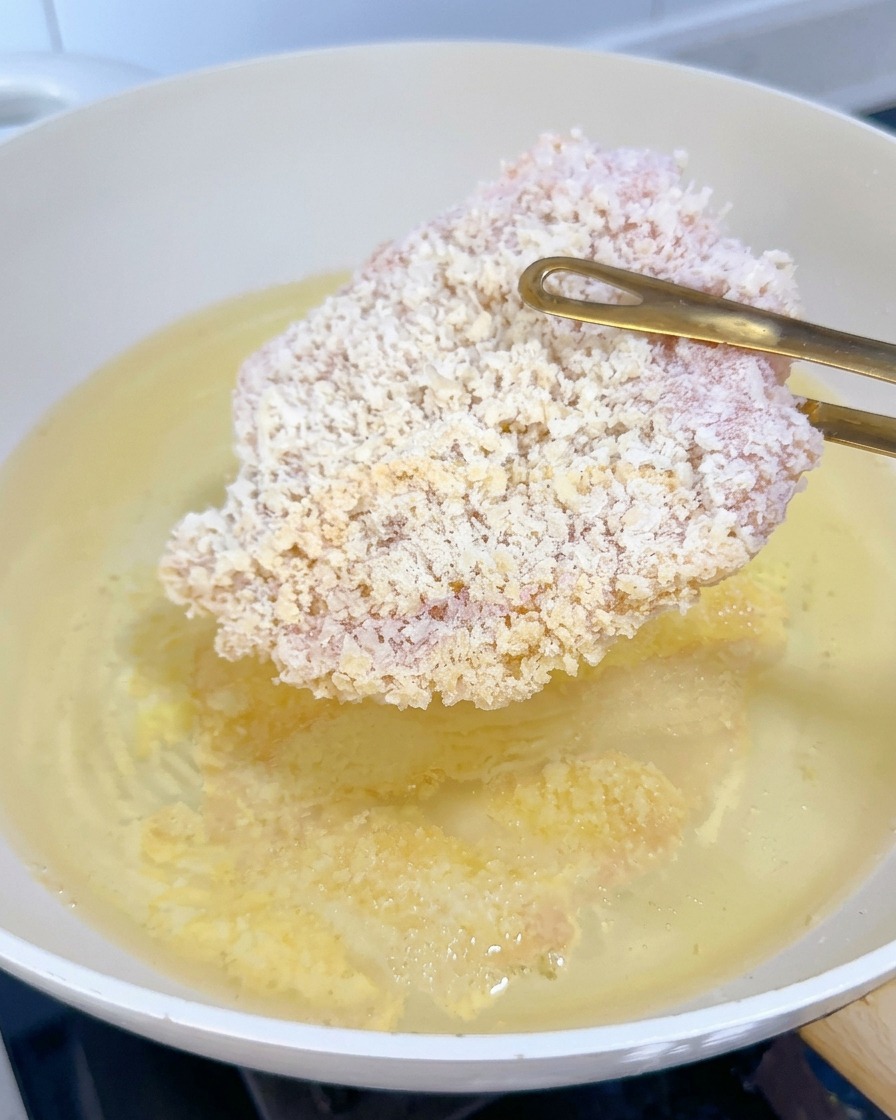 Una cotoletta di pollo panata ancora surgelata che viene immersa nellolio bollente con delle pinze.