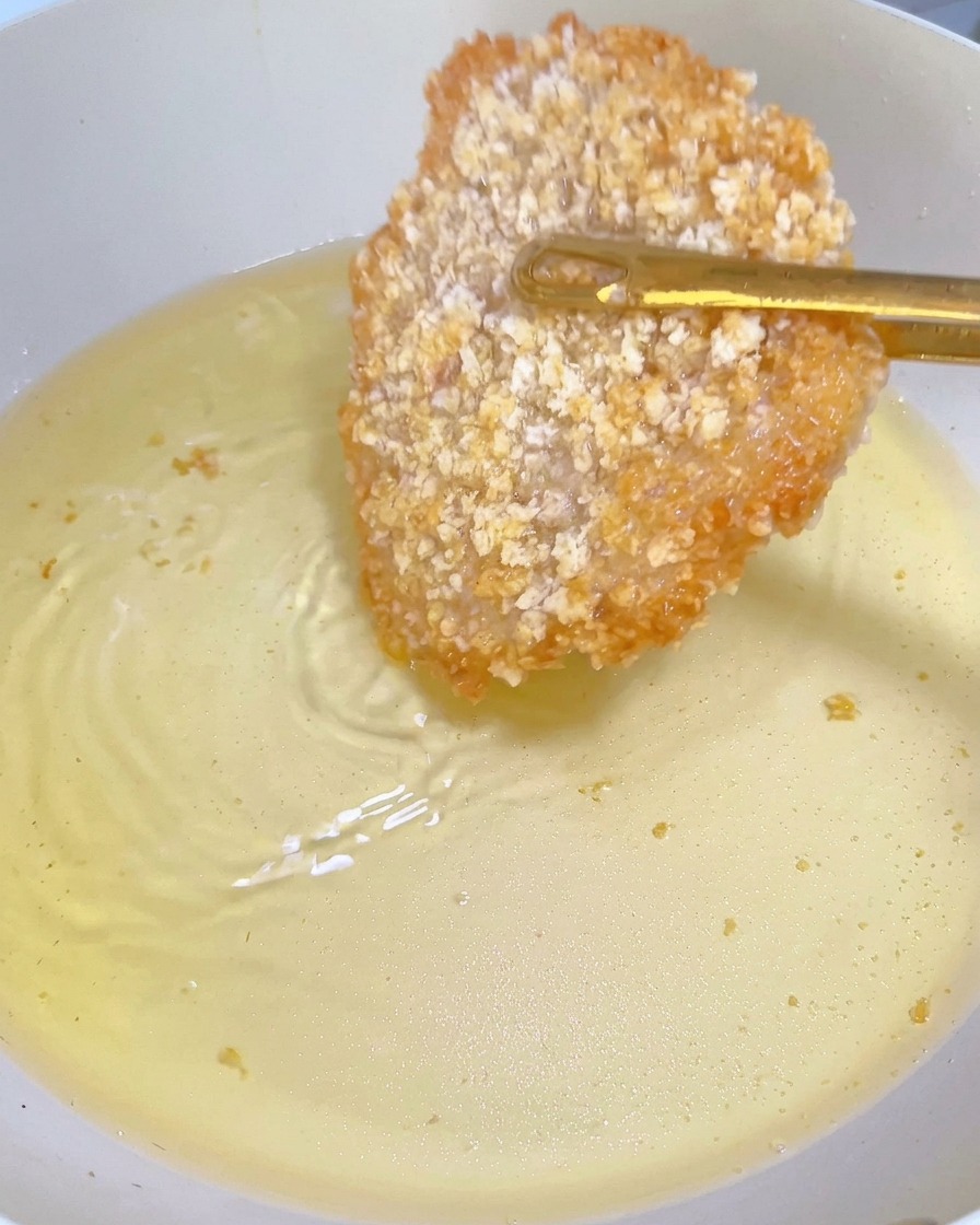 Una cotoletta di pollo parzialmente fritta con una crosticina dorata, girata con pinze in padella.