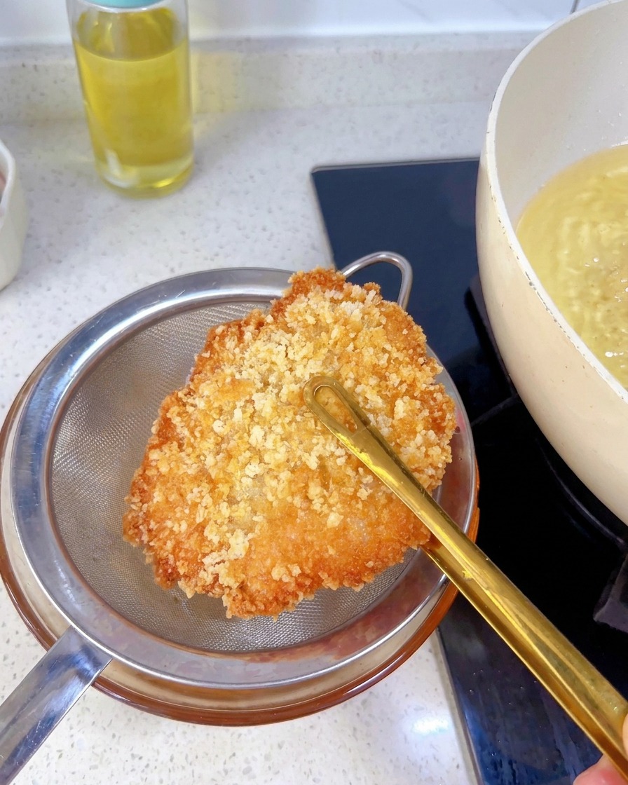 Una cotoletta di pollo dorata tenuta con le pinze sopra un colino di metallo per scolare lolio in eccesso.