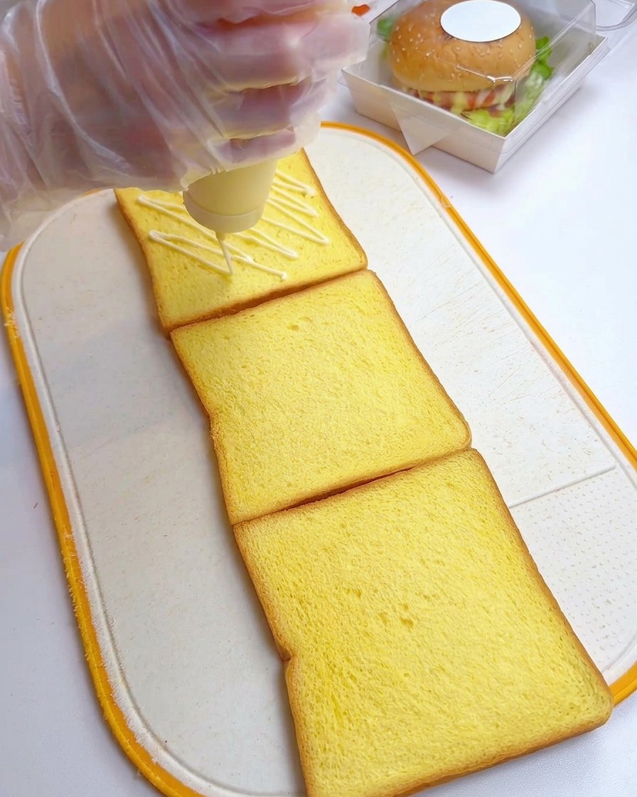 Una mano che versa maionese a zig-zag su tre fette di pane per toast su un tagliere.