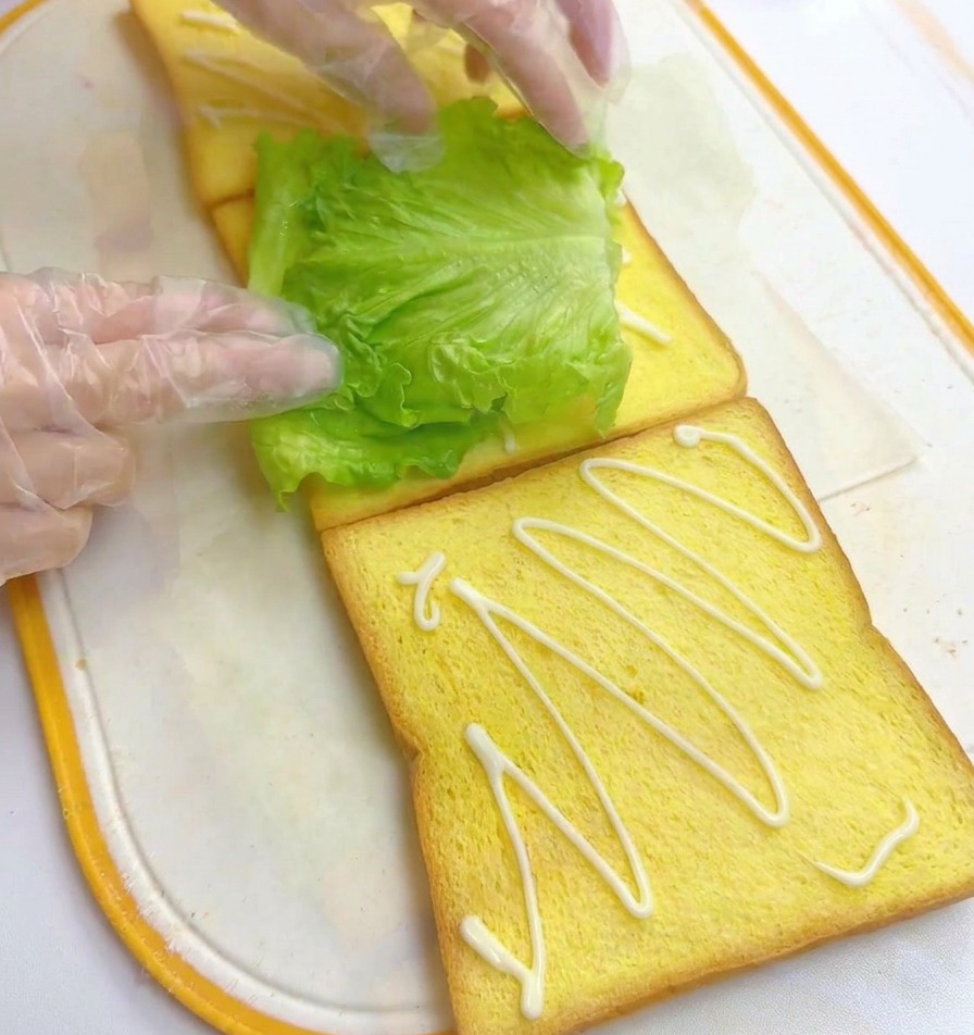 Mani che posizionano una foglia di lattuga fresca su una fetta di pane già condita con maionese.