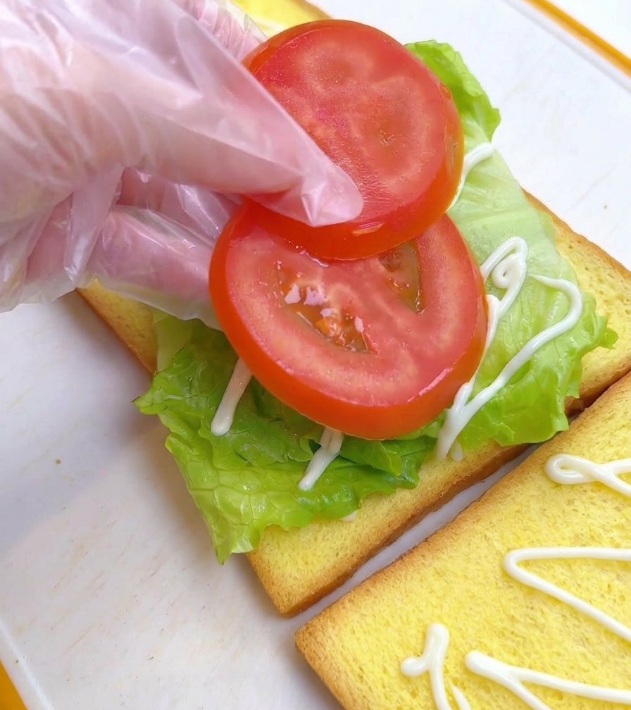 Dita che posizionano due fette tonde di pomodoro sopra la lattuga su una fetta di pane per toast.