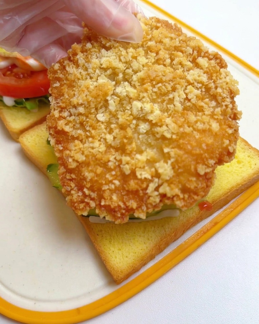 Una mano con guanto in plastica che posiziona la cotoletta di pollo fritta sopra i cetrioli sul pane.