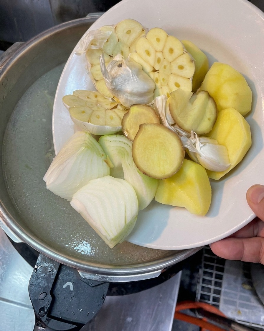 Un piatto con teste daglio tagliate a metà, cipolle tritate, fette di zenzero e pezzi di patata sopra una pentola di brodo bollente.