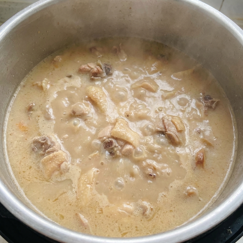 Una pentola con brodo tonkotsu bianco latte, denso e opaco, che bolle vigorosamente con pezzi di carne visibili nel liquido.