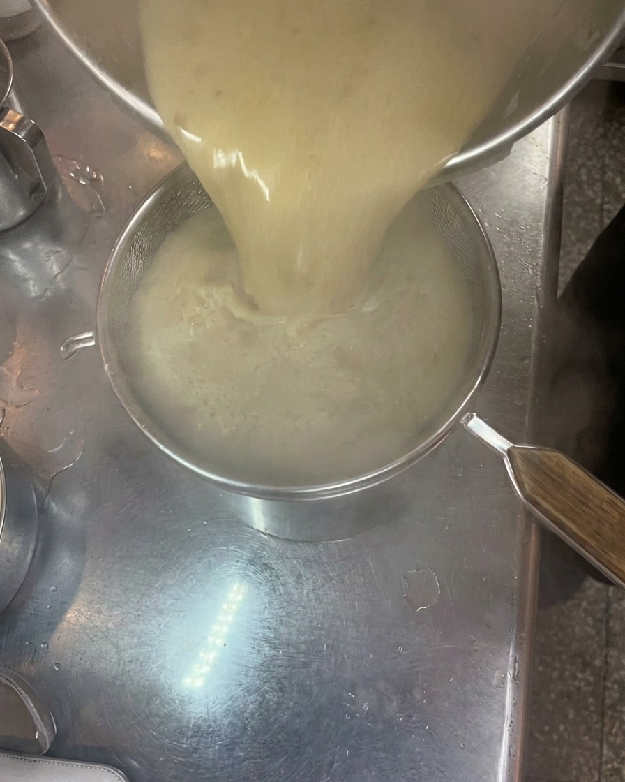Brodo tonkotsu denso e cremoso versato da una pentola attraverso un colino a maglia fine in unaltra pentola.