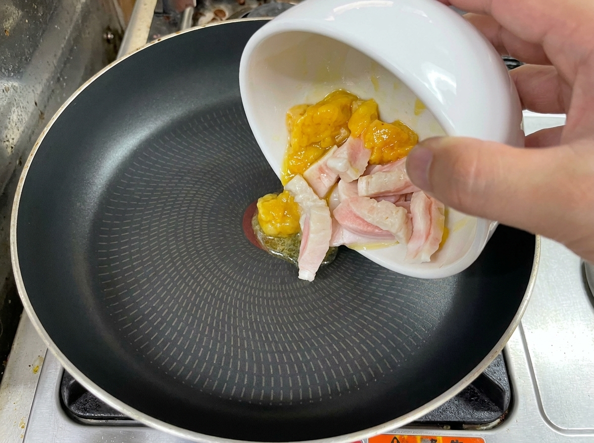 Una mano che versa pezzi di grasso crudo di pollo e maiale da una ciotola bianca in una padella nera.