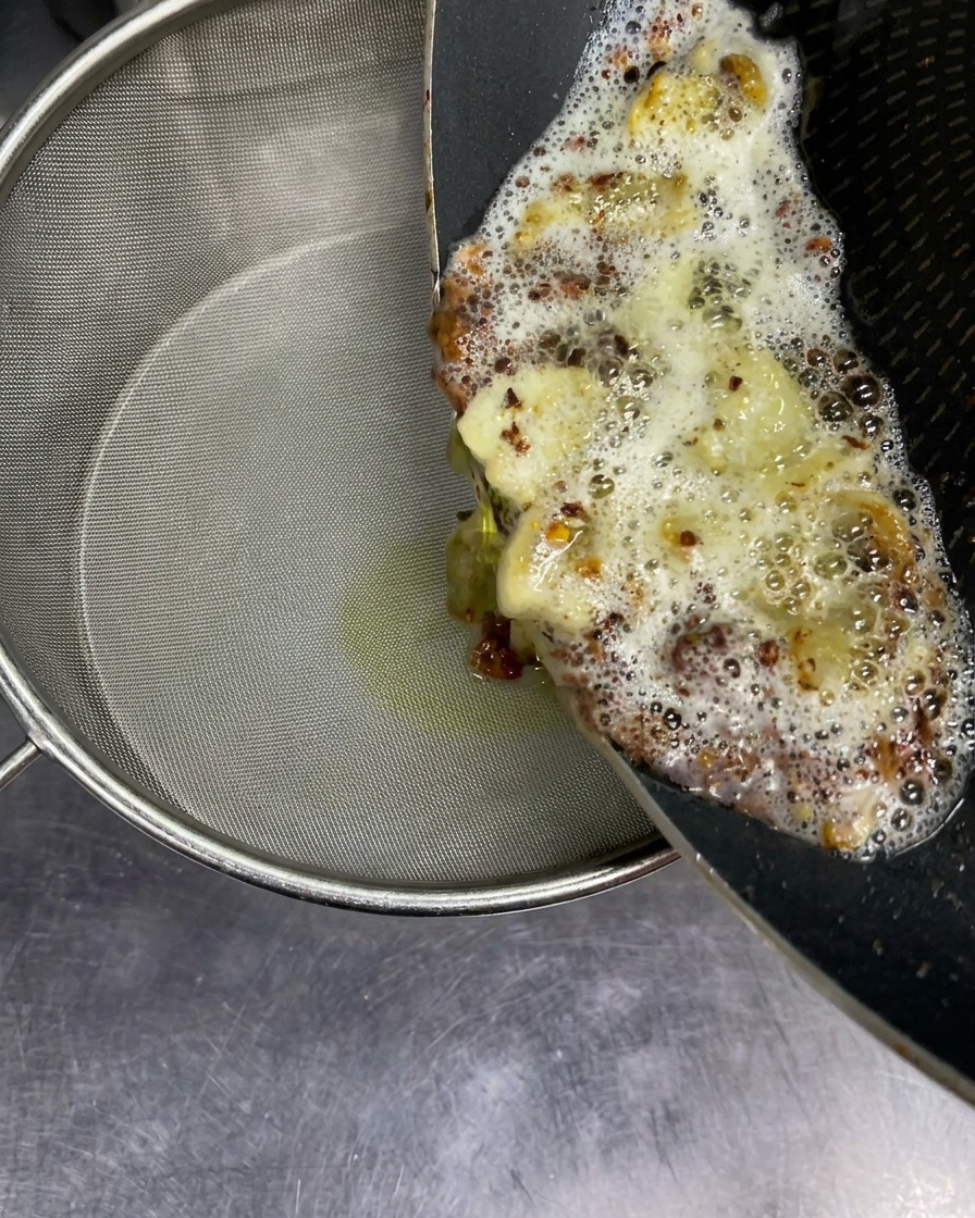 Olio aromatico bollente con pezzi di aglio fritto versato da una padella in un setaccio a maglia fine.