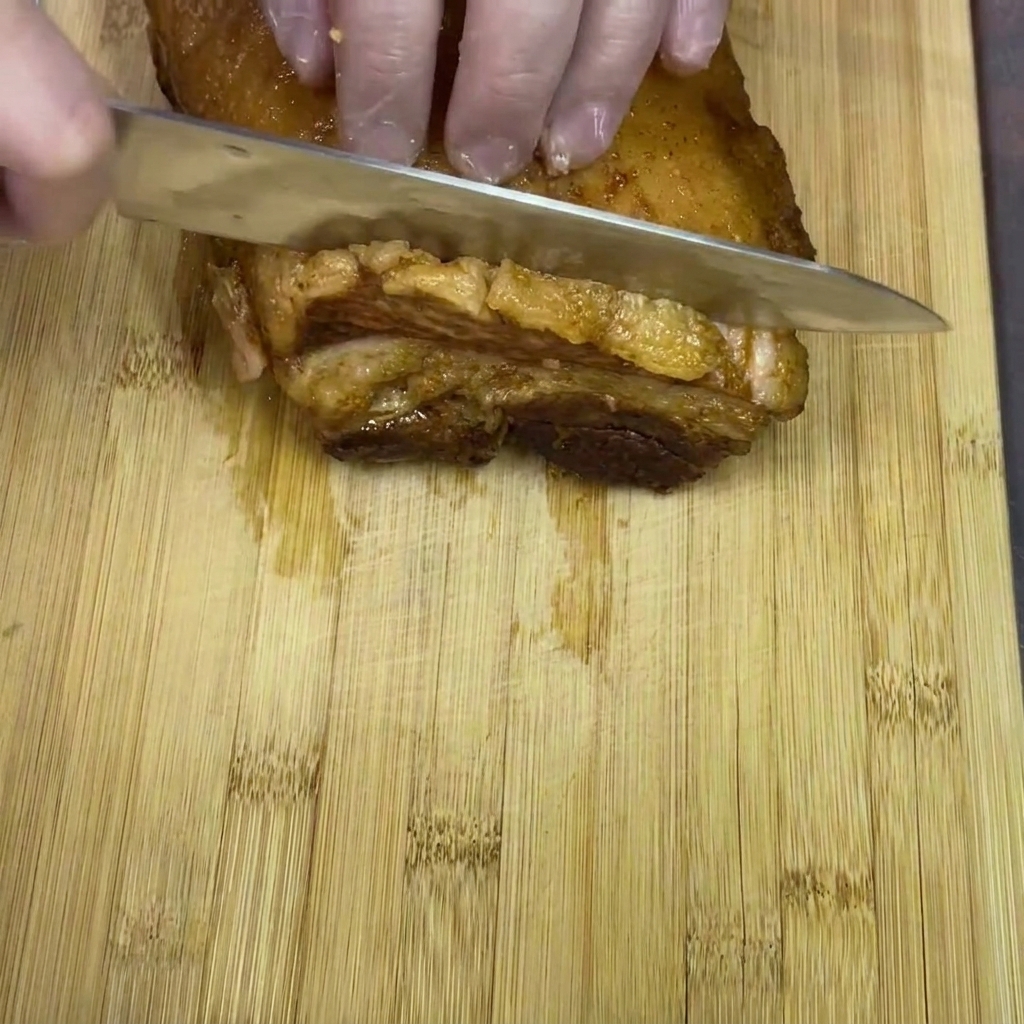 Mani con guanti che affettano un blocco di pancia di maiale brasata in pezzi spessi su un tagliere di legno.