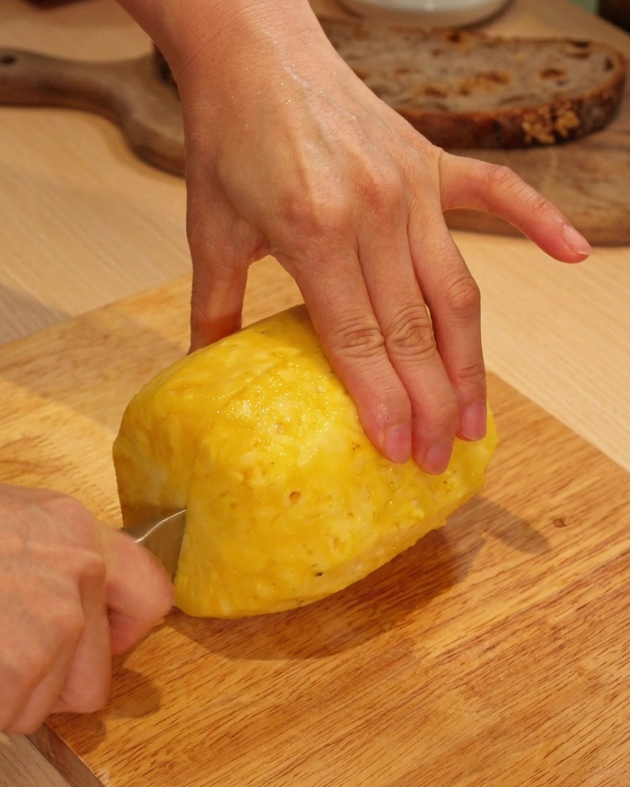 Mani che usano un coltello per affettare un ananas fresco sbucciato su un tagliere di legno.