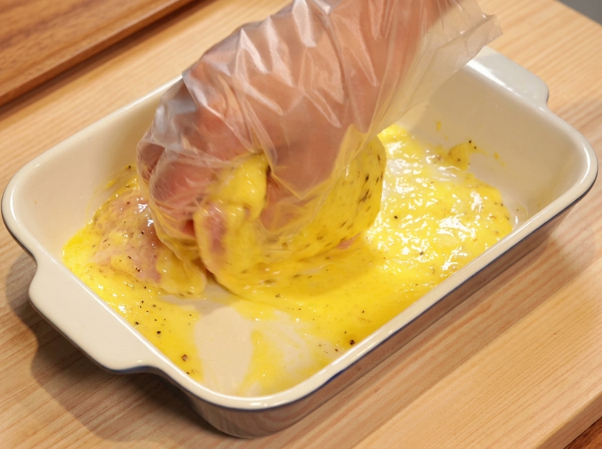 Una mano con guanto trasparente che massaggia il pollo nella marinatura gialla allananas.