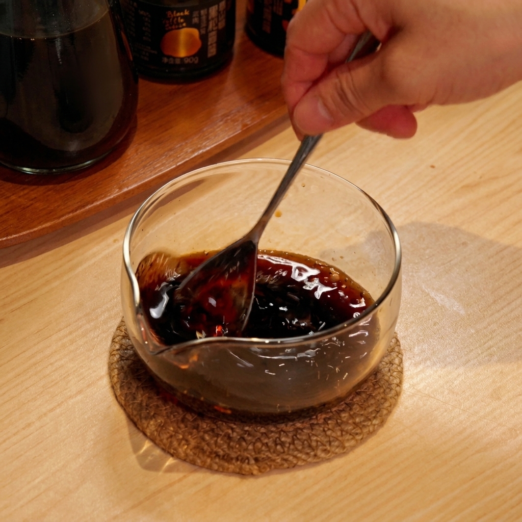 Una mano che usa un cucchiaio per mescolare una salsa scura in una piccola ciotola di vetro.