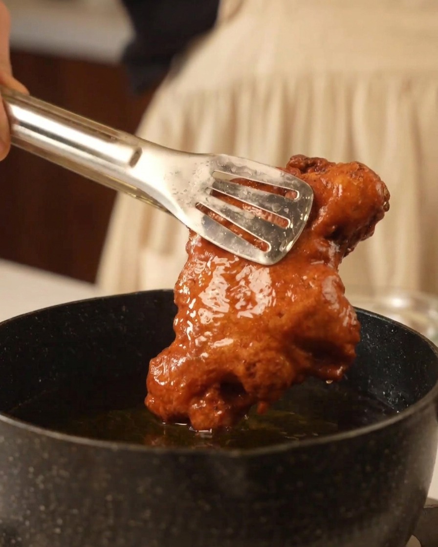Pinze di metallo che sollevano pollo fritto dorato e croccante dallolio bollente.