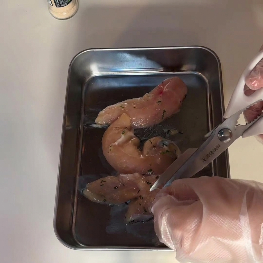 Mani con guanti che tagliano fette di petto di pollo crudo su un vassoio di acciaio.