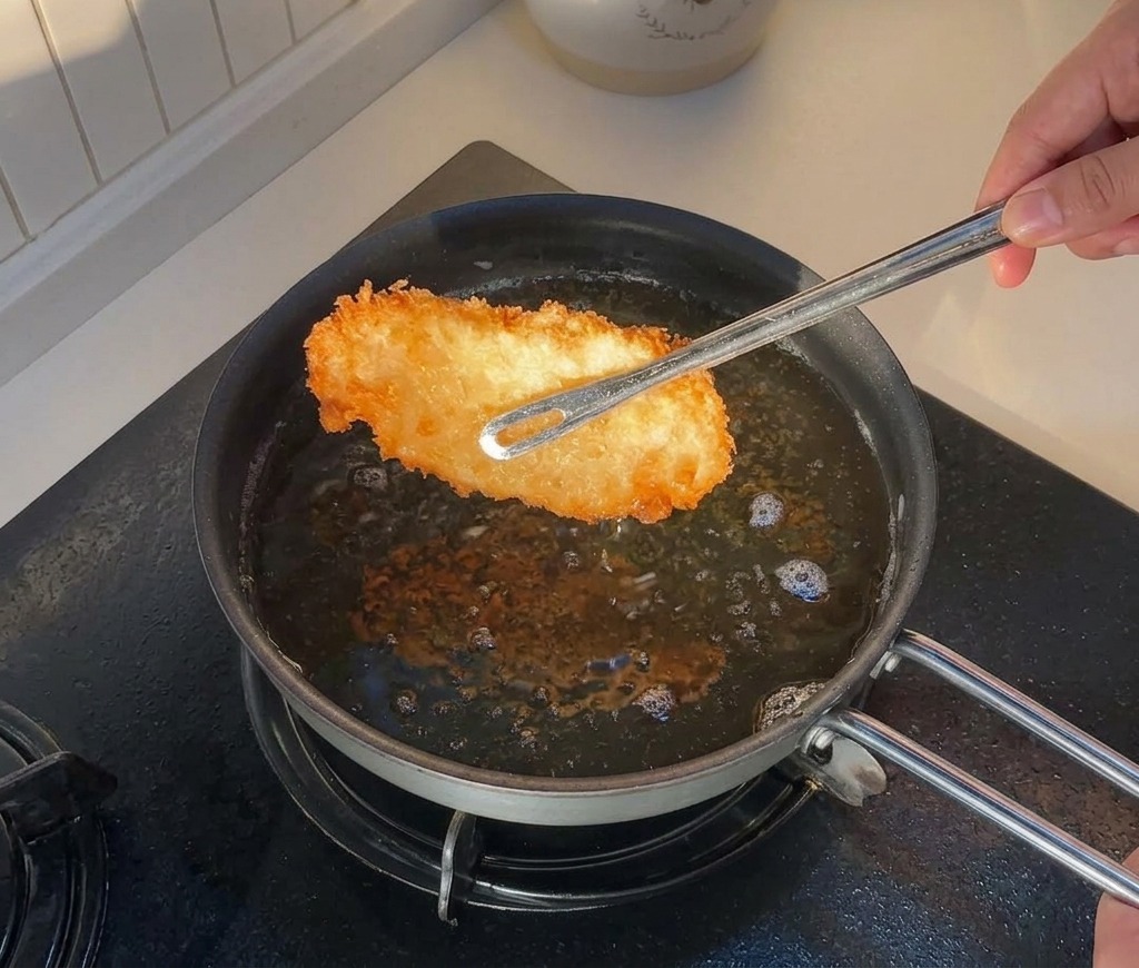 Pinze che sollevano un pezzo di pollo dorato da una padella con olio caldo.