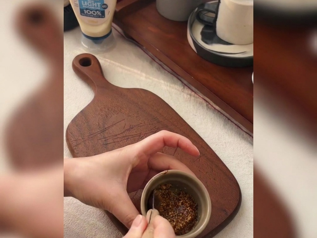 Mani che mescolano senape in grani e miele in una ciotolina di ceramica su un tagliere.
