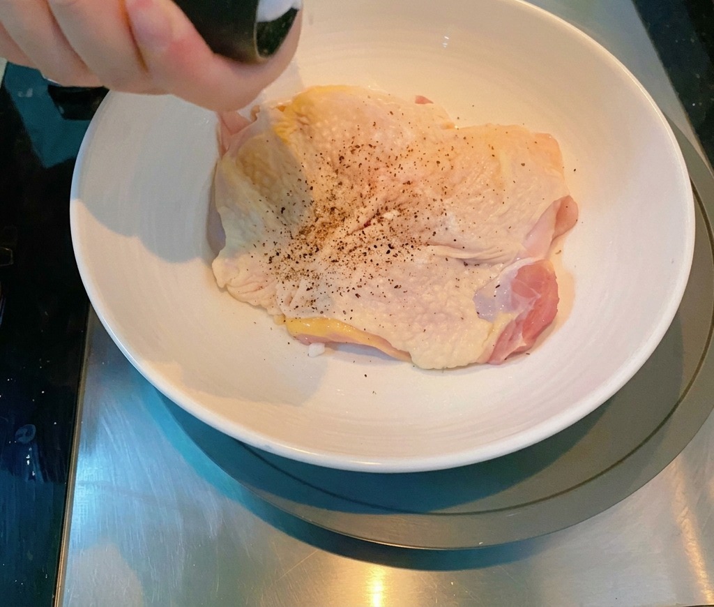 Una mano che usa un macinapepe per condire una coscia di pollo cruda in una ciotola di ceramica.