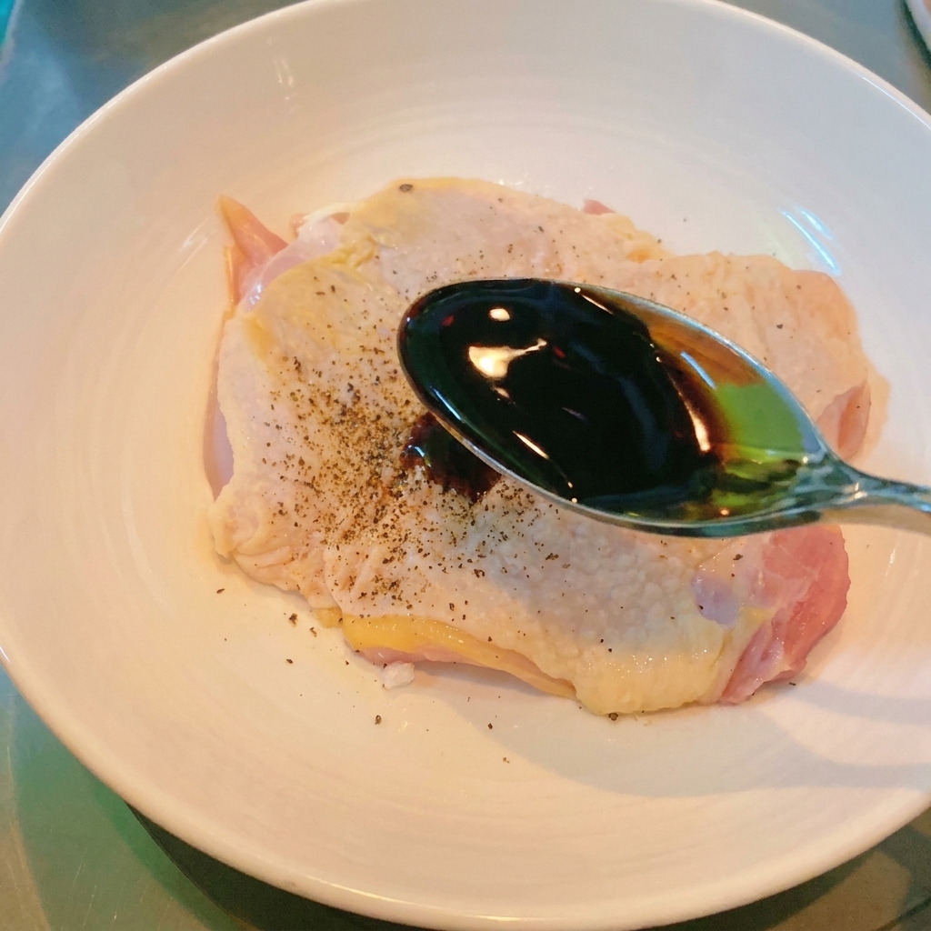 Un cucchiaio di metallo che versa salsa di soia scura su un pezzo di pollo crudo in una ciotola.
