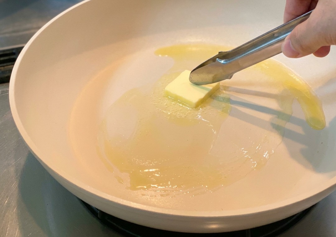 Un pezzetto di burro spostato con pinze in una padella mentre inizia a sciogliersi.