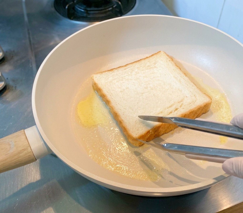 Una fetta di pane per sandwich tostata in padella con burro fuso, tenuta ferma con le pinze.