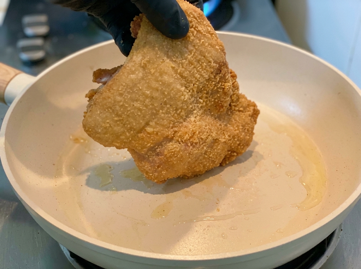 Una mano con guanto che mette la coscia di pollo infarinata in una padella calda.