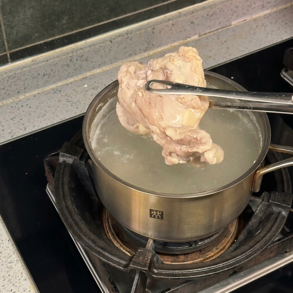 Un pezzo di pollo sollevato con pinze metalliche sopra una pentola dacciaio con acqua in ebollizione.