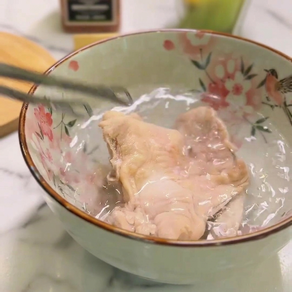 Pollo cotto immerso in una ciotola di ceramica con acqua fredda, tenuto con pinze metalliche.