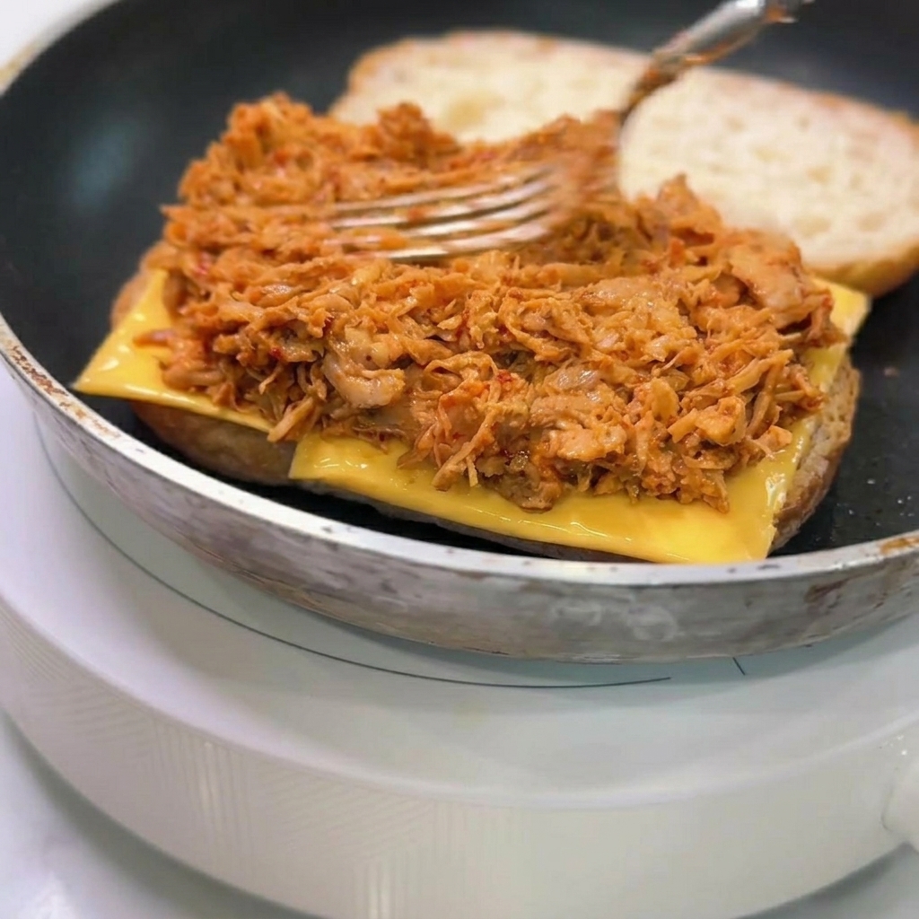 Una forchetta che stende uno strato generoso di pollo sfilacciato in salsa arancione sul formaggio e la ciabatta.