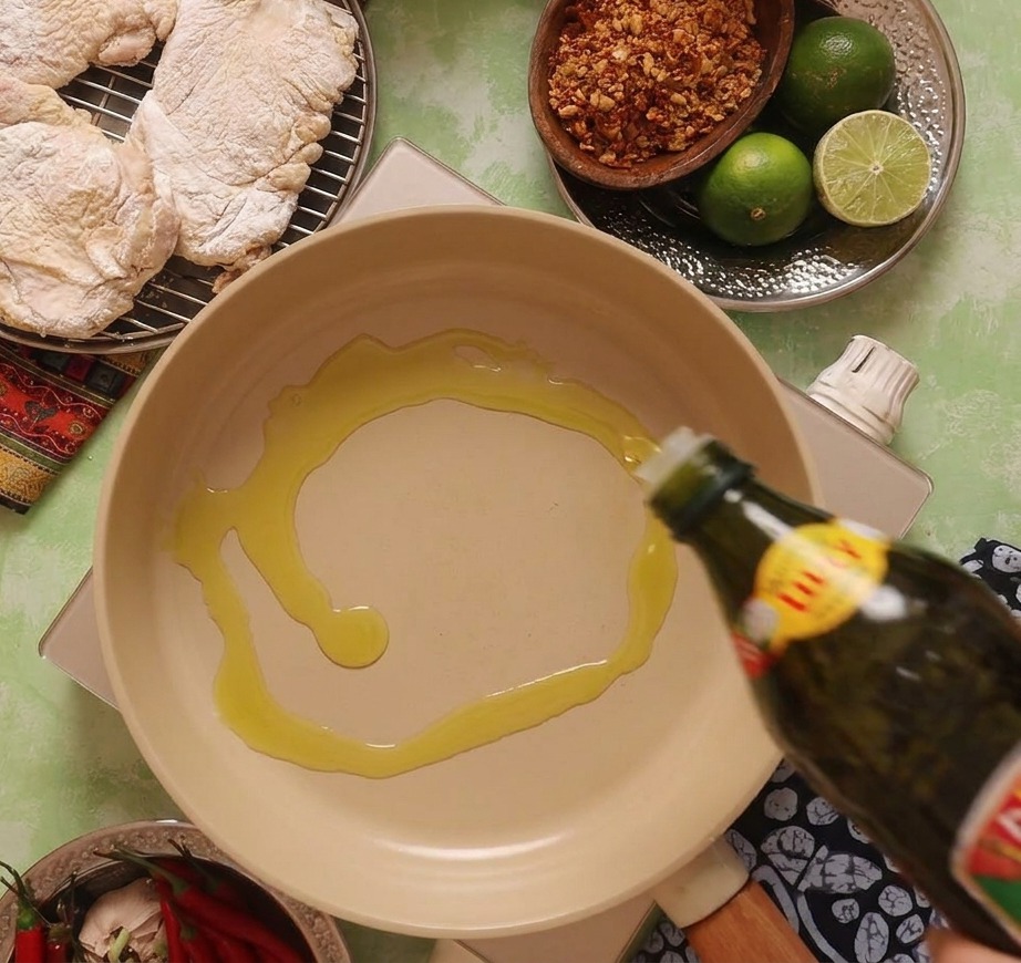Olio doliva versato da una bottiglia scura in una padella di ceramica beige su un fornello portatile.
