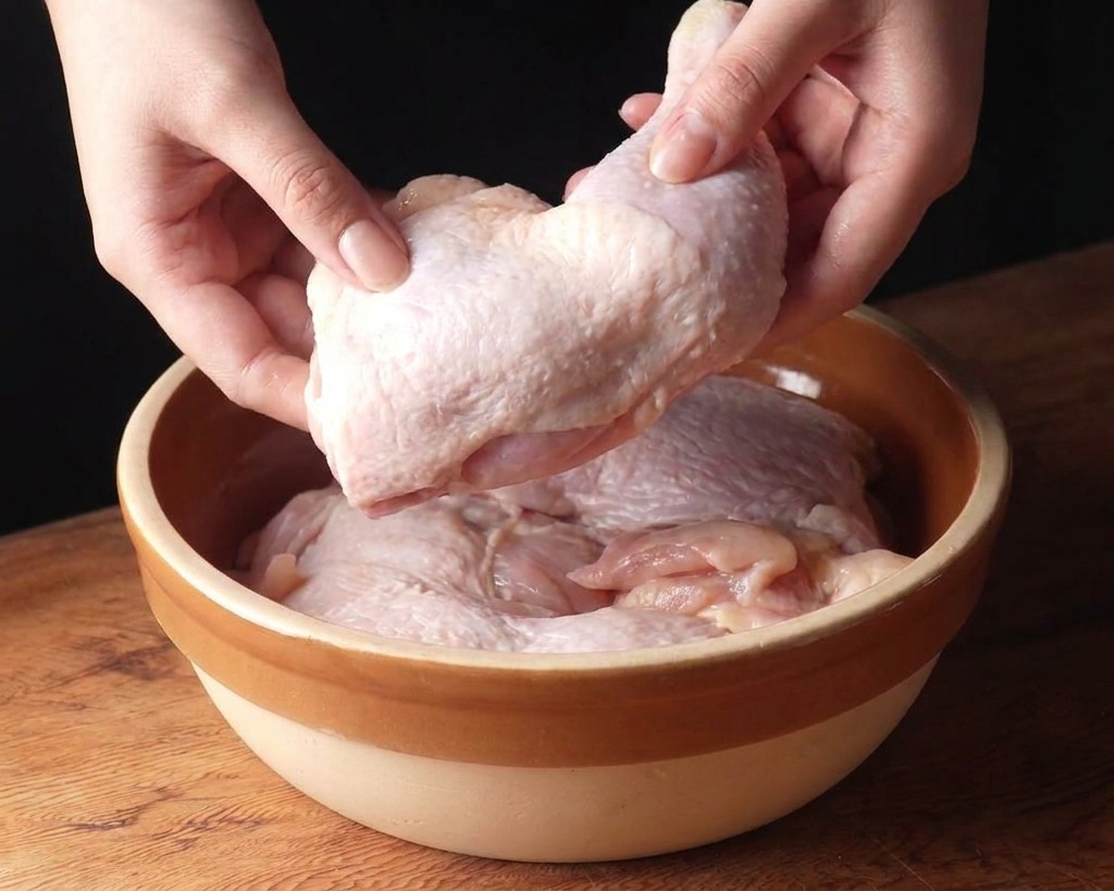 Mani che adagiano sovracosce di pollo crudo in una ciotola di ceramica beige su un piano di legno.