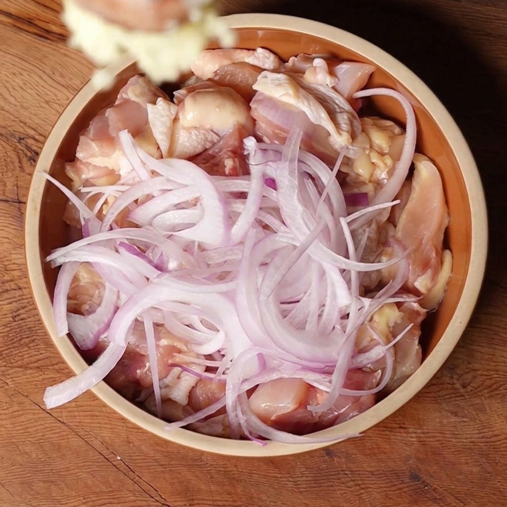 Aglio tritato aggiunto in una ciotola con pezzi di pollo crudo e cipolla rossa affettata.